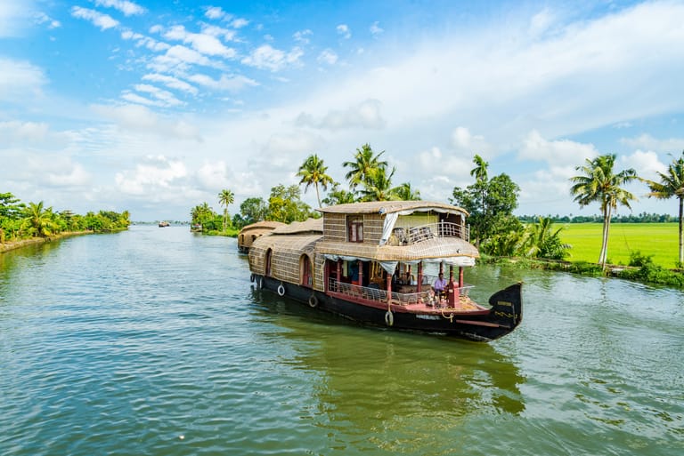 A traditional houseboat with distinctive woven bamboo or cane roofing cruises through the serene backwaters surrounded by lush tropical vegetation. The vessel