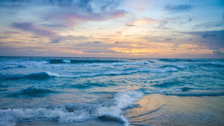 A serene coastal seascape captured during the golden hour, featuring gentle ocean waves rolling onto a sandy beach at sunset or sunrise. The image evokes a