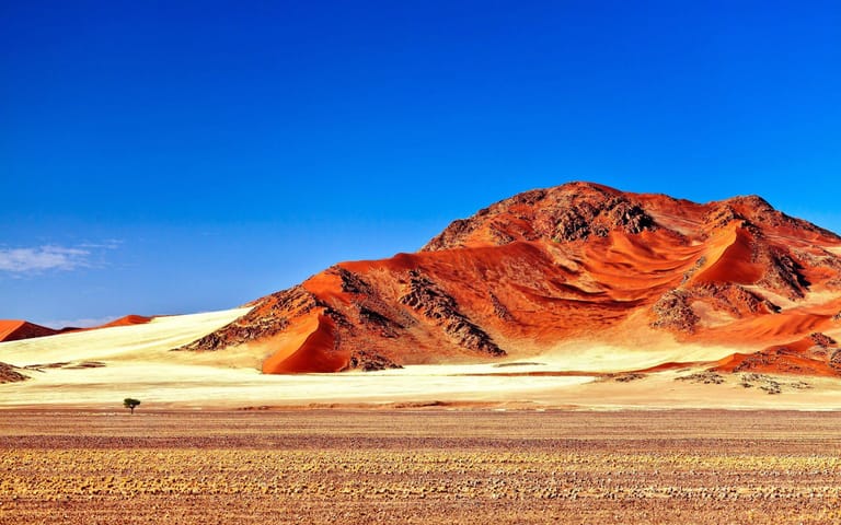 A breathtaking desert landscape showcasing dramatic red and orange sand dunes against towering rocky mountains under a brilliant blue sky. The scene captures
