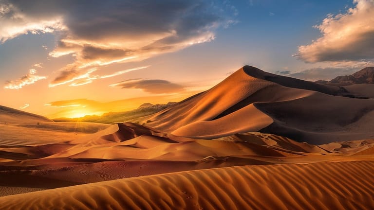 A breathtaking desert landscape featuring majestic sand dunes illuminated by golden sunset light. The image captures the dramatic interplay of light and shadow