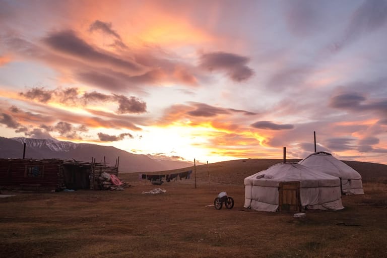 A traditional Mongolian ger (yurt) stands in a remote pastoral landscape during a dramatic sunset. The circular nomadic dwelling with its distinctive white
